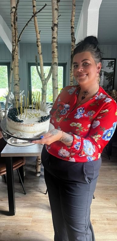 Eine Frau in einem bunten Pullover hält einen Kuchen mit Kerzen in der Hand.