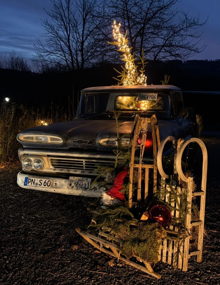 Alter Pick-up mit Weihnachtsdekoration, umgeben von festlicher Beleuchtung und Tannenzweigen.