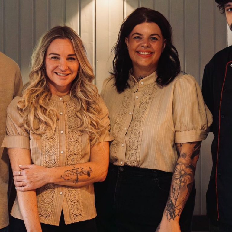 Drei Personen lächeln freundlich in eleganter Kleidung vor einem hellen Hintergrund.