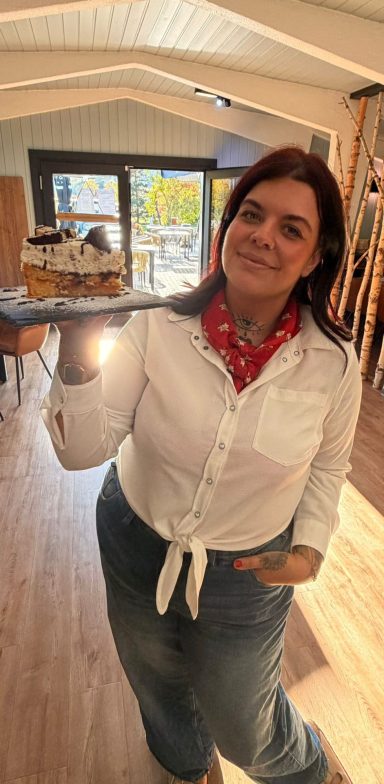 Frau mit rotem Schal präsentiert einen Kuchen auf einem Tablett in einem lichtdurchfluteten Raum.